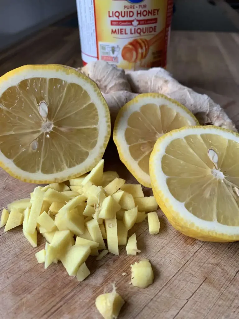 On a wooden cutting board is a jar of honey with some fresh ginger and a sliced up lemon. in the very front some sliced up ginger