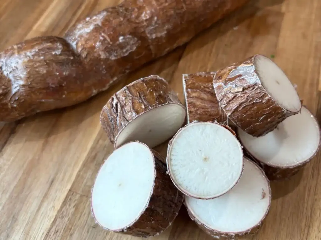 whole cassava in the back with sliced, unpeeled cassava slices in the front. All lying on a wooden cutting board