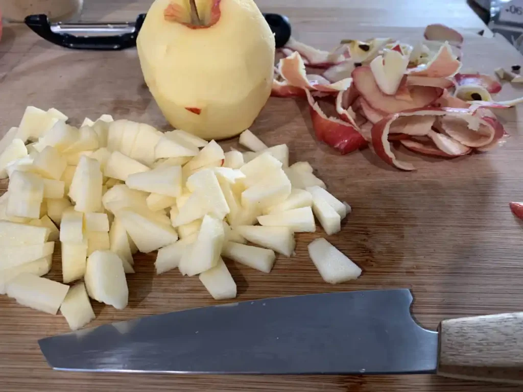 On a wooden cutting board lies a peeled apple as well as a diced apple. In the front a sharp kitchen knife and in the back a vegetable peeler