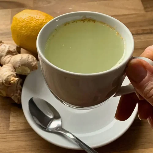 hand holding a white cup containing a soothing lemon ginger and honey drink sitting above a saucer and a teaspoon. Standing on a wooden counter with some fresh ginger and a lemon next to it