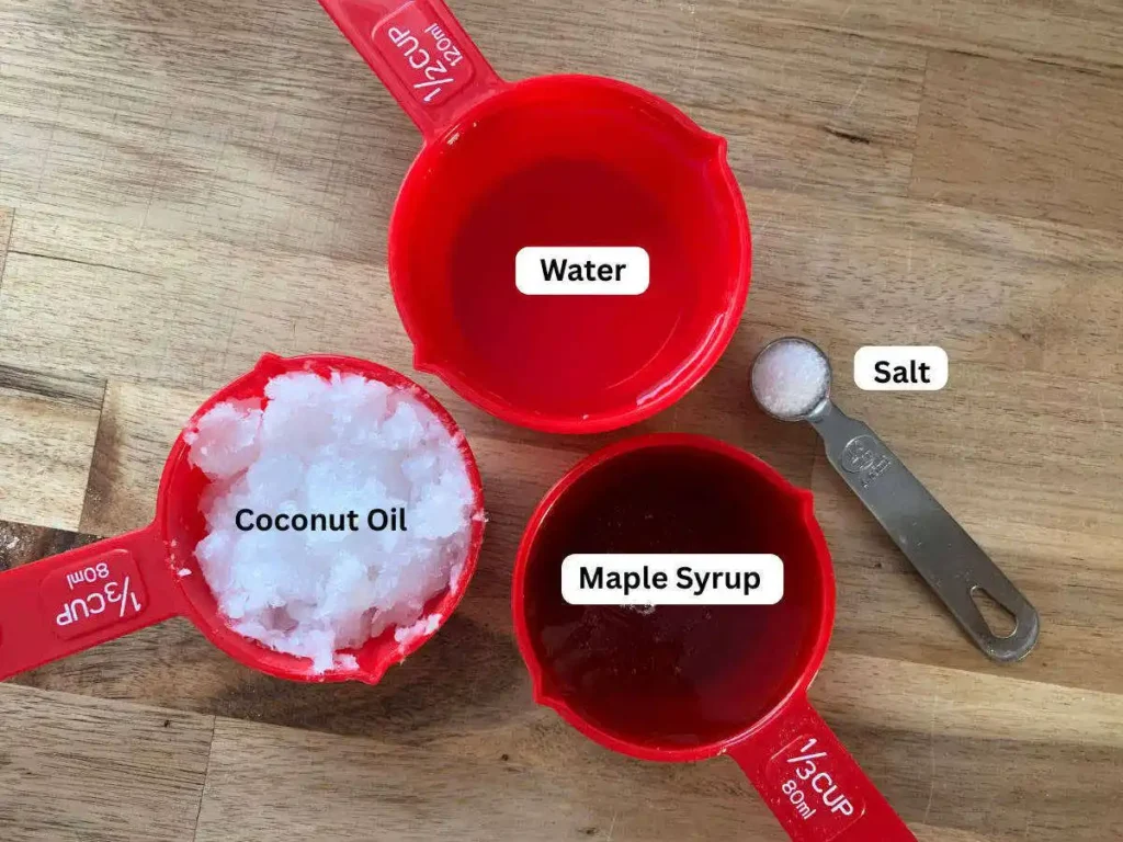 Wet ingredeints for apple muffins sitting on a wooden counter top. In red measuring cups the labels read water, maple syrup and coconut oil. In a silver measuring spoon is salt.