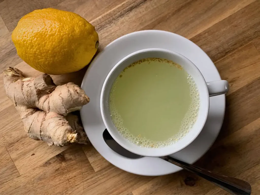 white cup containing a soothing lemon honey and ginger hot drink sitting on a saucer and a teaspoon. Standing on a wooden counter with some fresh ginger and a lemon next to it