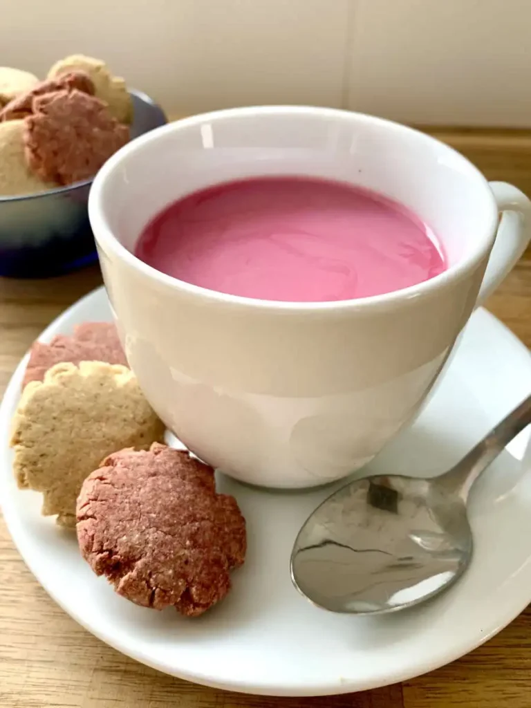 white cup with a pink drink staning on a white saucer. On the saucer is a teaspoon and three cookies (two pink and white). Behind is a blue glass bowl with more pink and white cookies