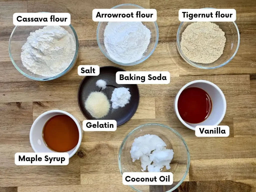 Ingredients for shortbread cookies in individual bowls sitting on a wooden counter. Ingredeints include cassava flour, arrowroot flur, tigernut flour, salt, baking soda, gelatin, maple syrup, vanilla, and coconut oil