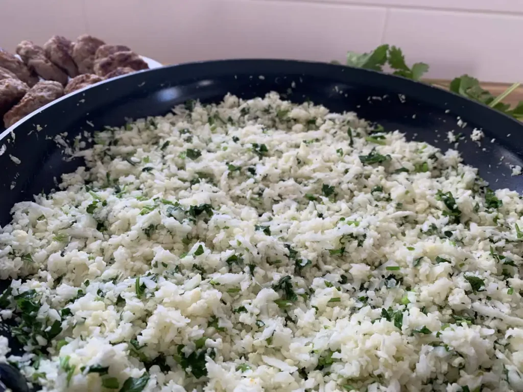 Cauliflower rice with fresh lime and cilantro in a large frying pan with fresh cilantro and a bowl of pork sausages in the background