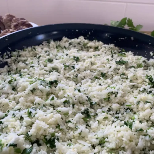 Cauliflower rice with fresh lime and cilantro in a large frying pan with fresh cilantro and a bowl of pork sausages in the background