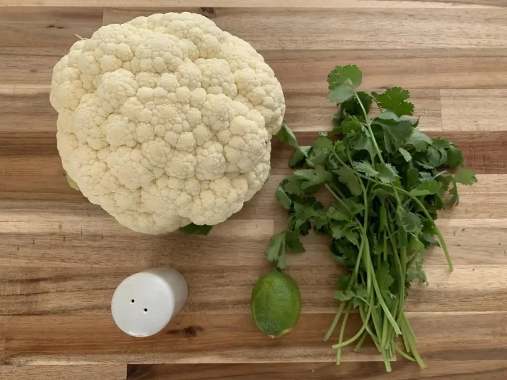 On a wooden cutting board are the ingredients for AIP cauliflower rice, which includes salt, lime, cilantro and cauliflower.